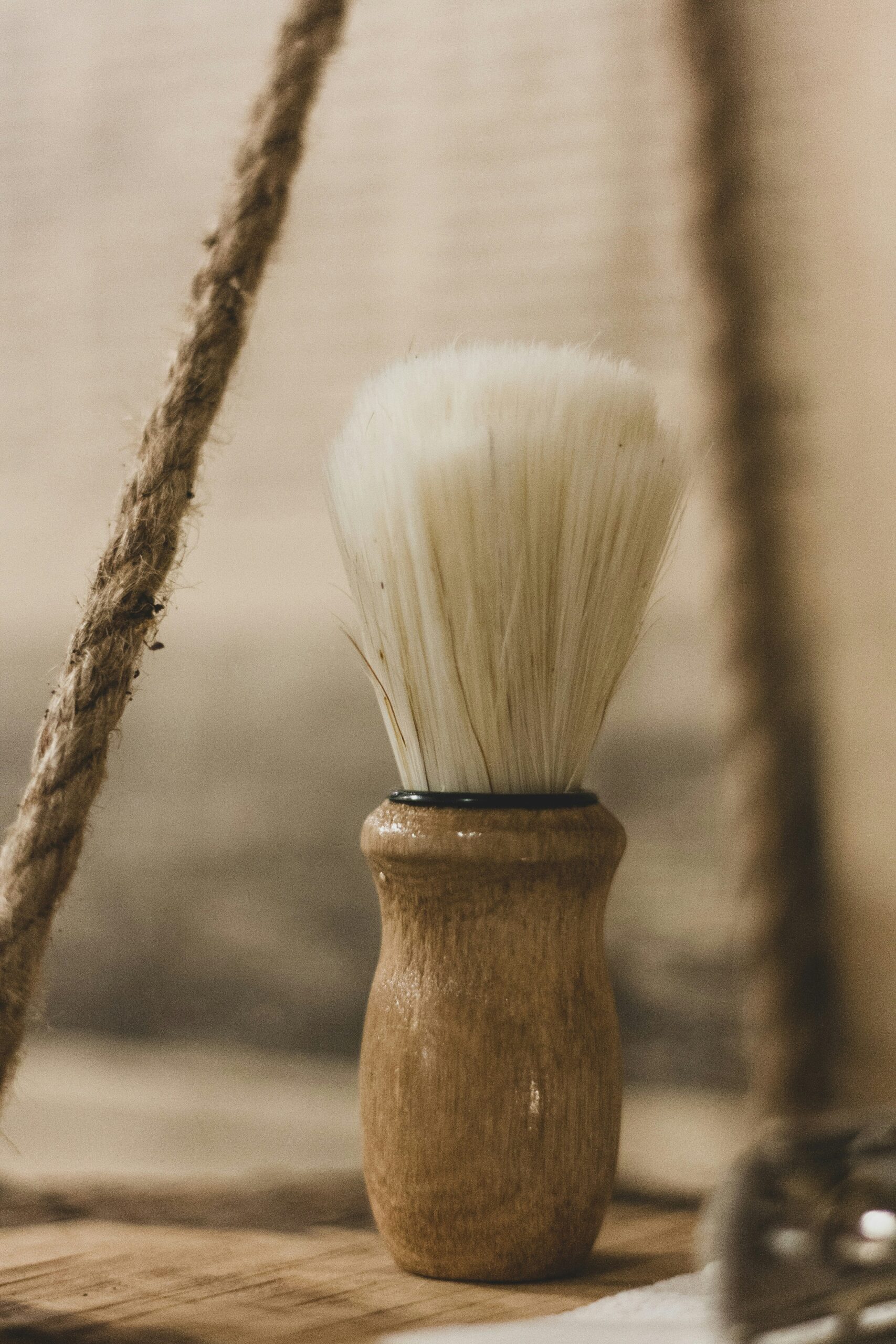 A close-up, rustic-style photograph of a traditional wooden-handled shaving brush with light-colored bristles, positioned next to a thick hanging rope.