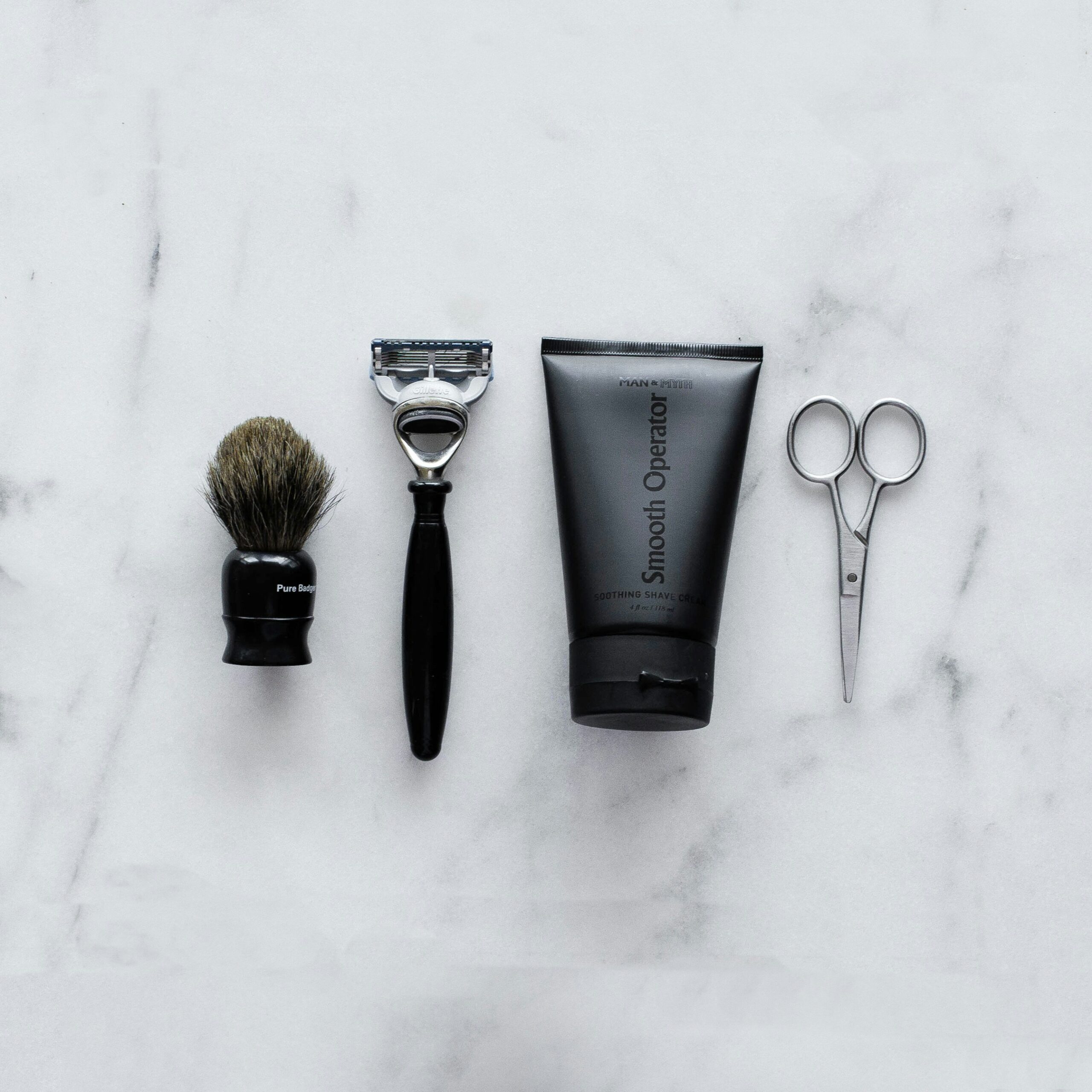 Top-down view of a minimalist men's grooming kit on white marble, including a black shaving brush, a modern razor, a black tube of "Smooth Operator" shave cream, and small silver grooming scissors.